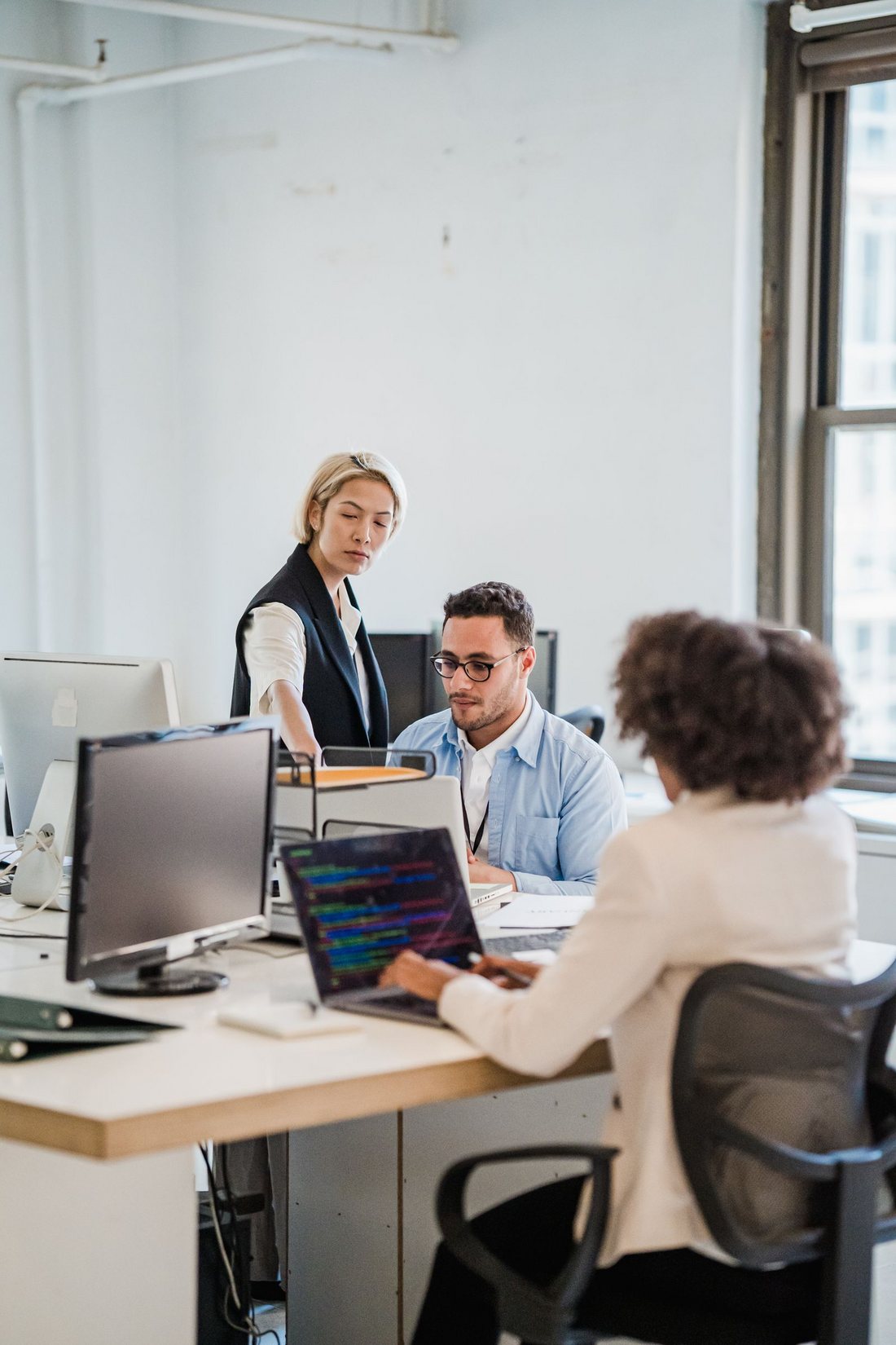 Three consultants collaborating around a desk in a bright office, reviewing code on a laptop during a working session
